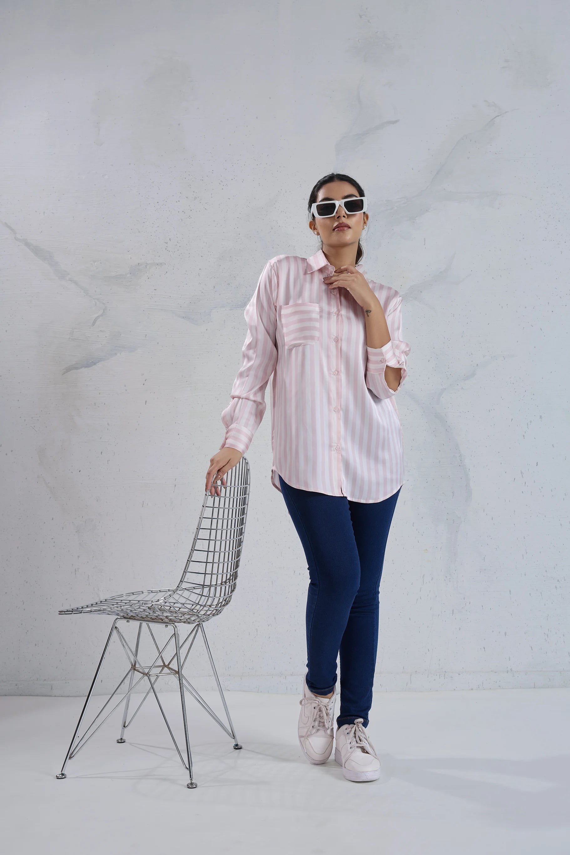 Person wearing a light pink shirt and dark blue jeans standing next to a wire chair against a white background