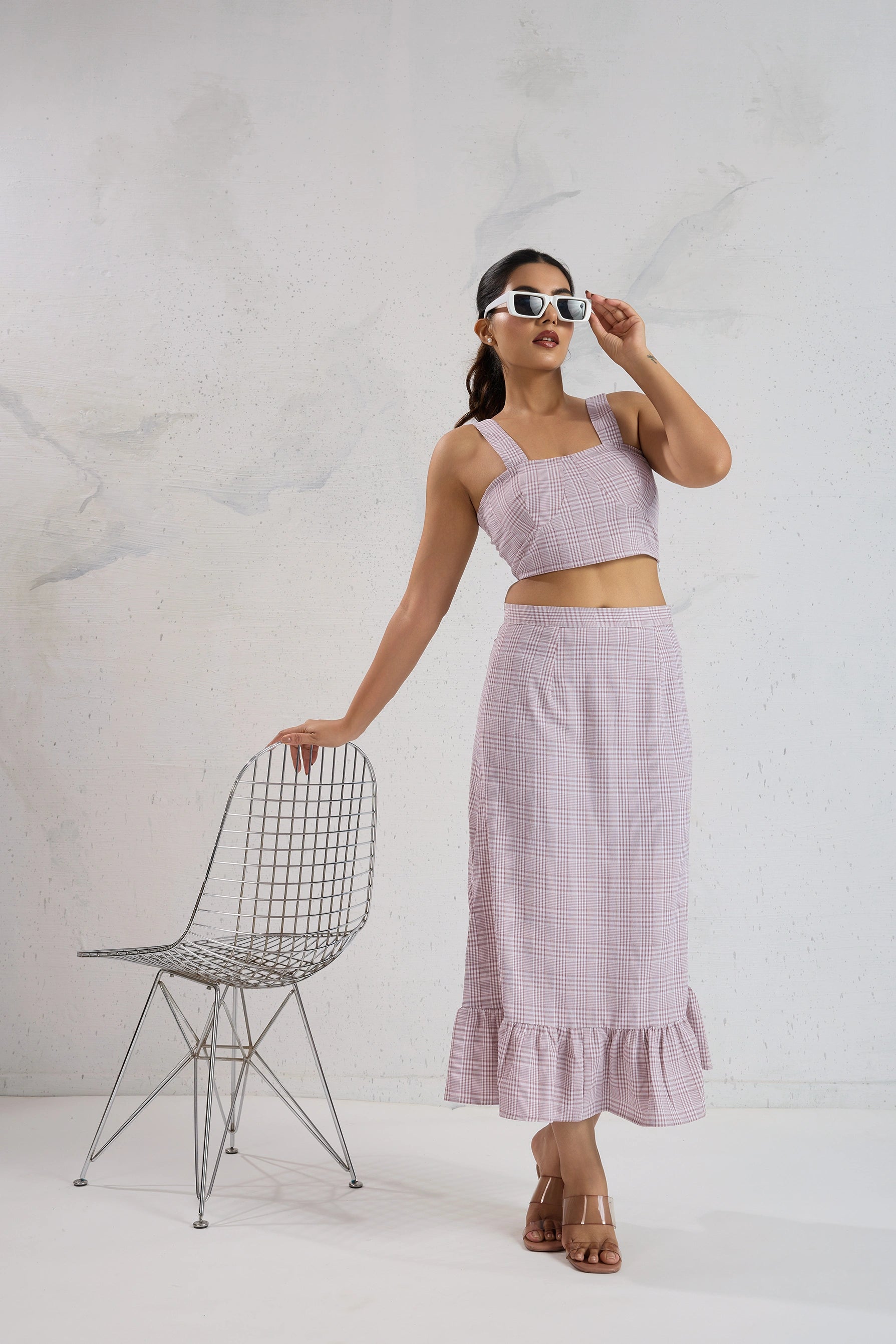 Woman wearing a matching pink top and skirt standing next to a wire chair against a white background