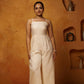 A woman standing against a textured background, wearing a beige linen jumpsuit with spaghetti straps and a tailored fit.