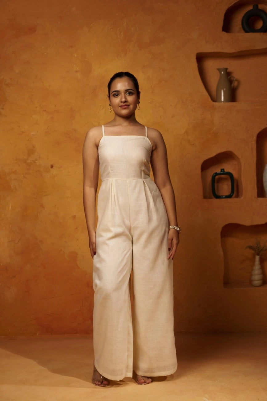 A woman standing against a textured background, wearing a beige linen jumpsuit with spaghetti straps and a tailored fit.