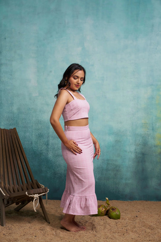 Woman in a pink outfit standing against a textured teal wall with a wooden chair and coconuts on the ground.