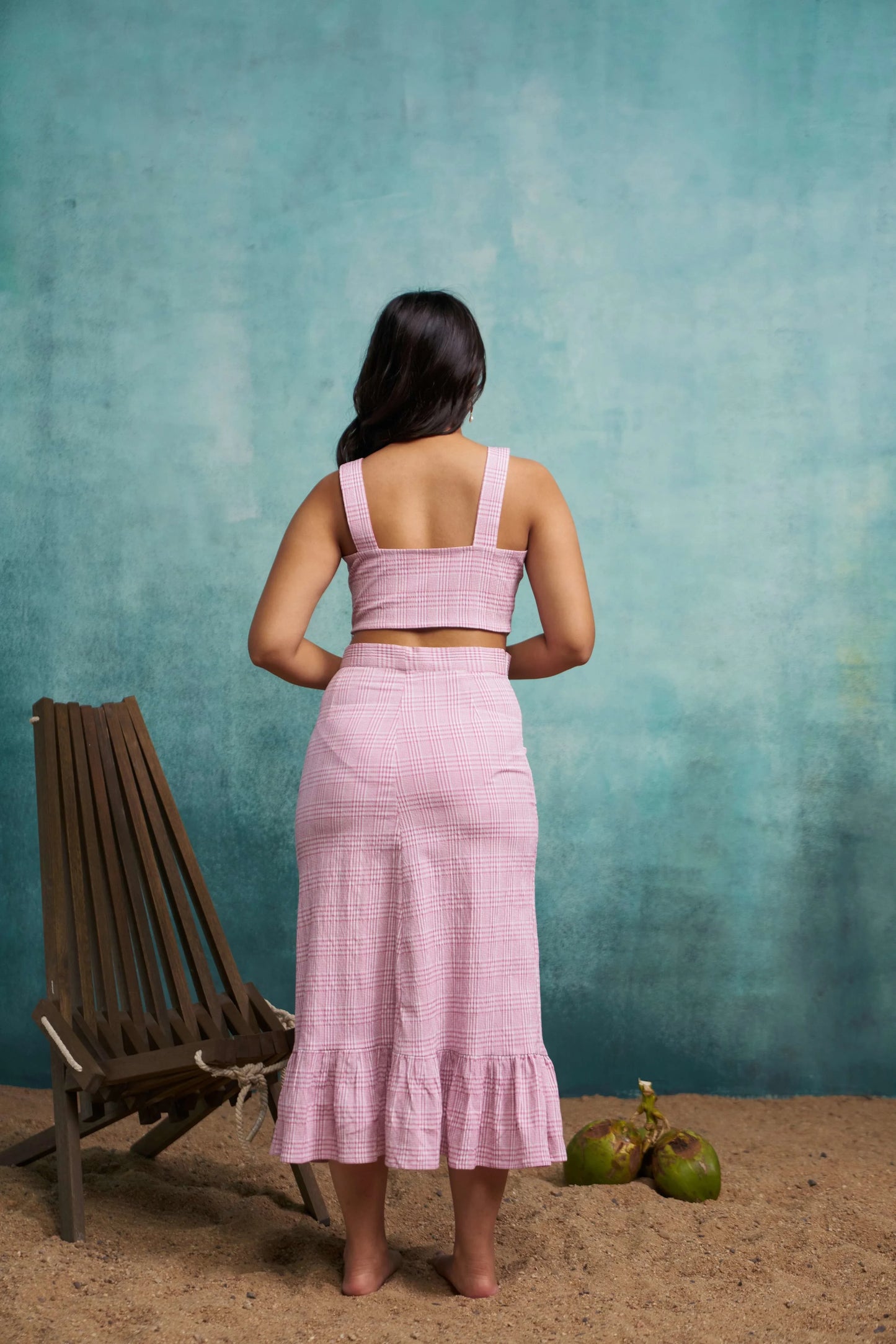 Woman in a pink outfit standing against a textured teal wall with a chair and fruits on the ground.