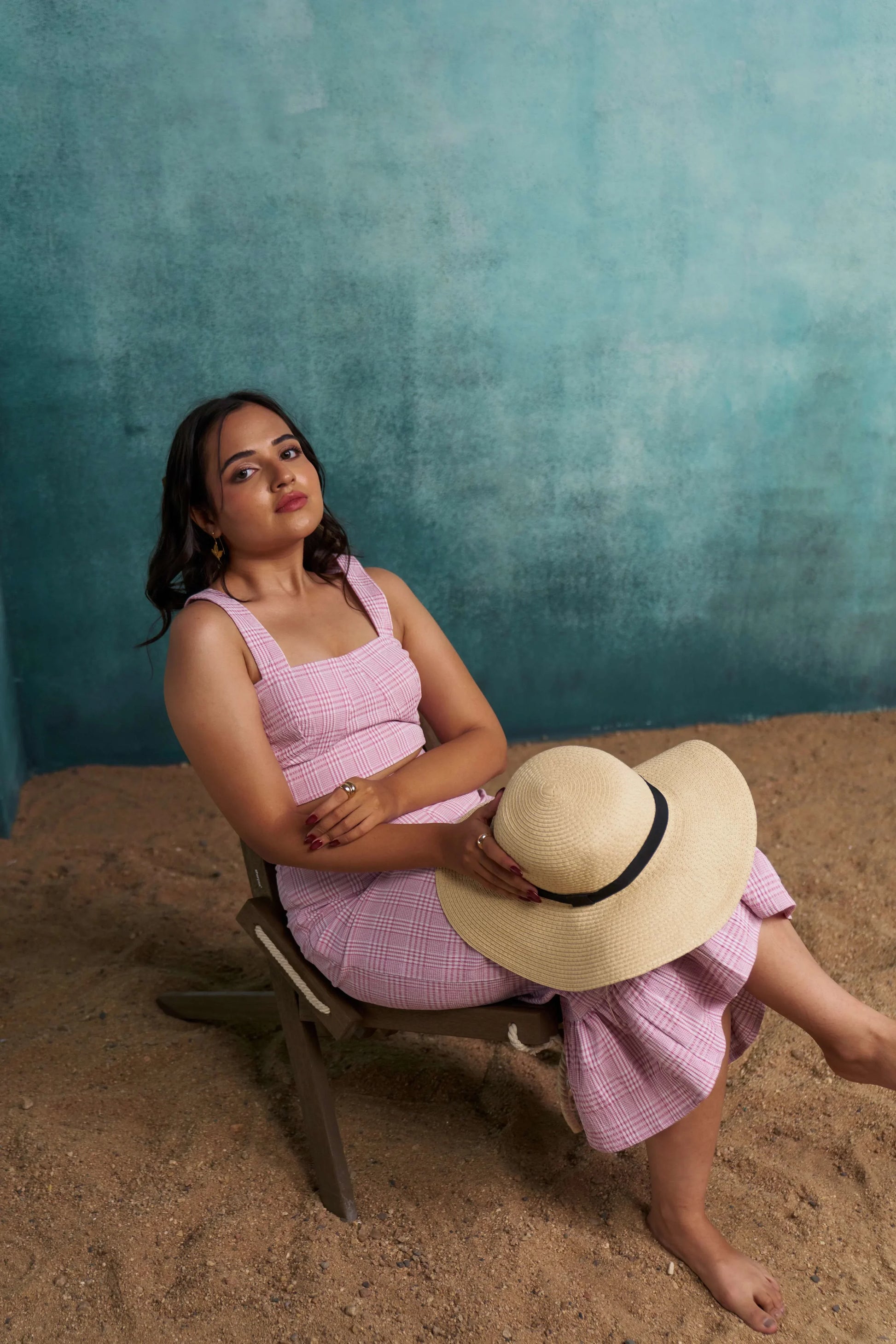 Woman in a pink dress sitting on sand with a straw hat, against a teal wall.