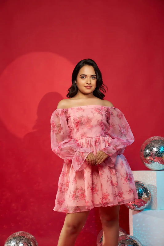 A woman standing in front of a red background wearing a pink floral off-shoulder organza dress with balloon sleeves.