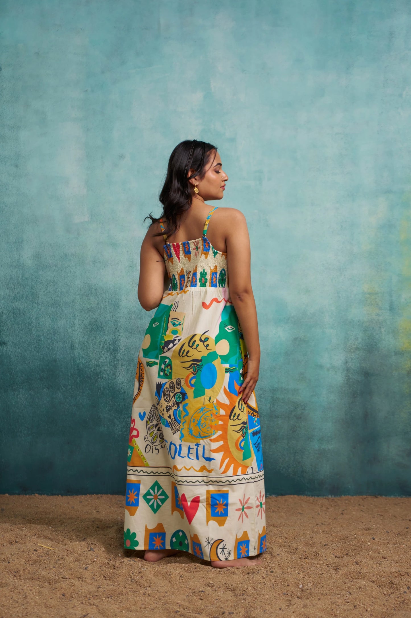 Woman wearing a colorful dress with a pattern, standing against a textured blue wall.