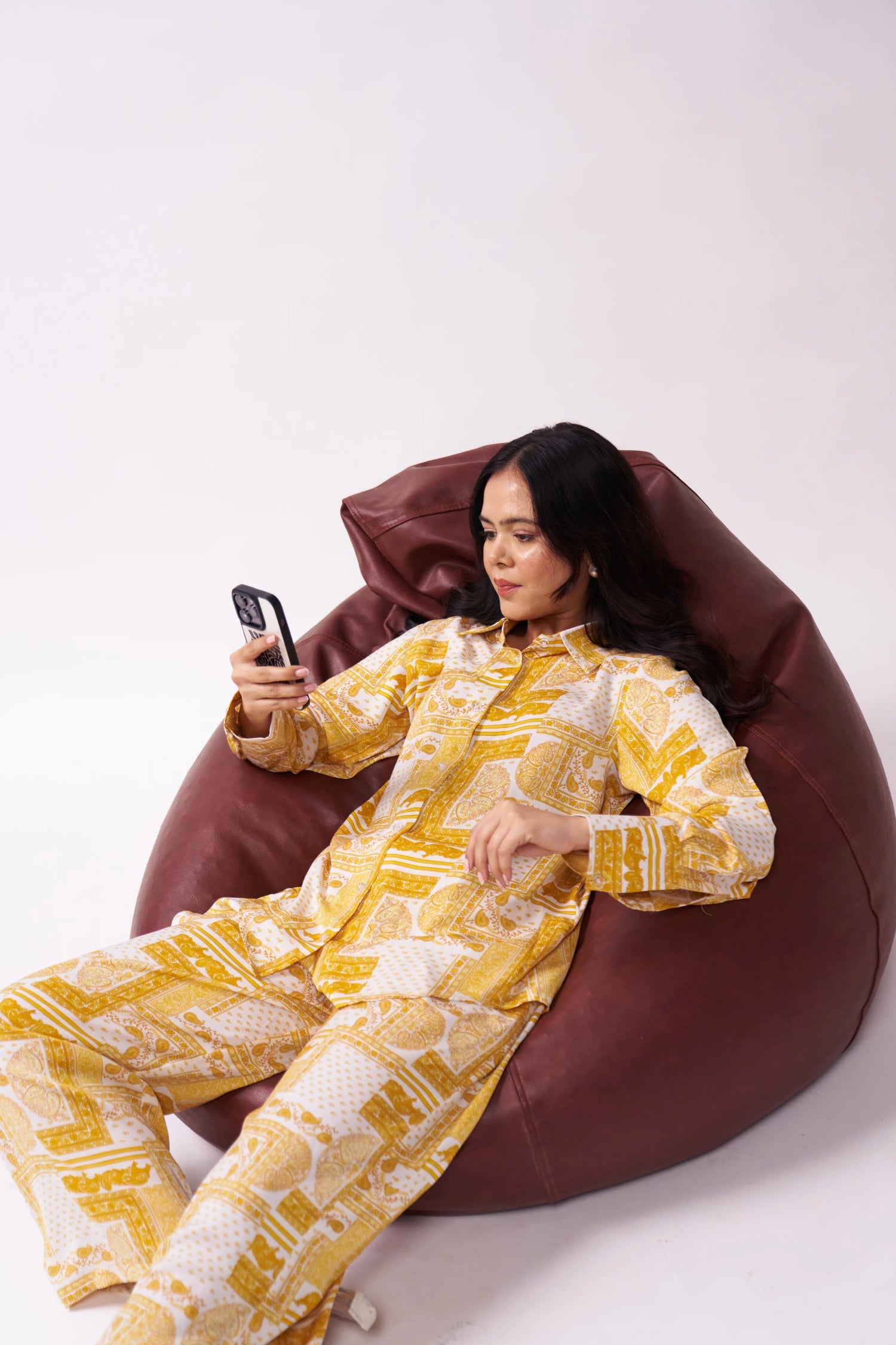Woman in yellow and white patterned outfit sitting on a brown bean bag chair, using a phone.