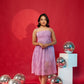 A woman standing in a studio setting with a red background, wearing a lavender pleated party dress with a sleeveless design.
