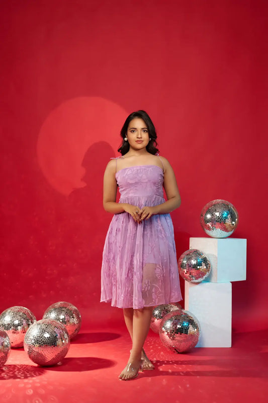A woman standing in a studio setting with a red background, wearing a lavender pleated party dress with a sleeveless design.