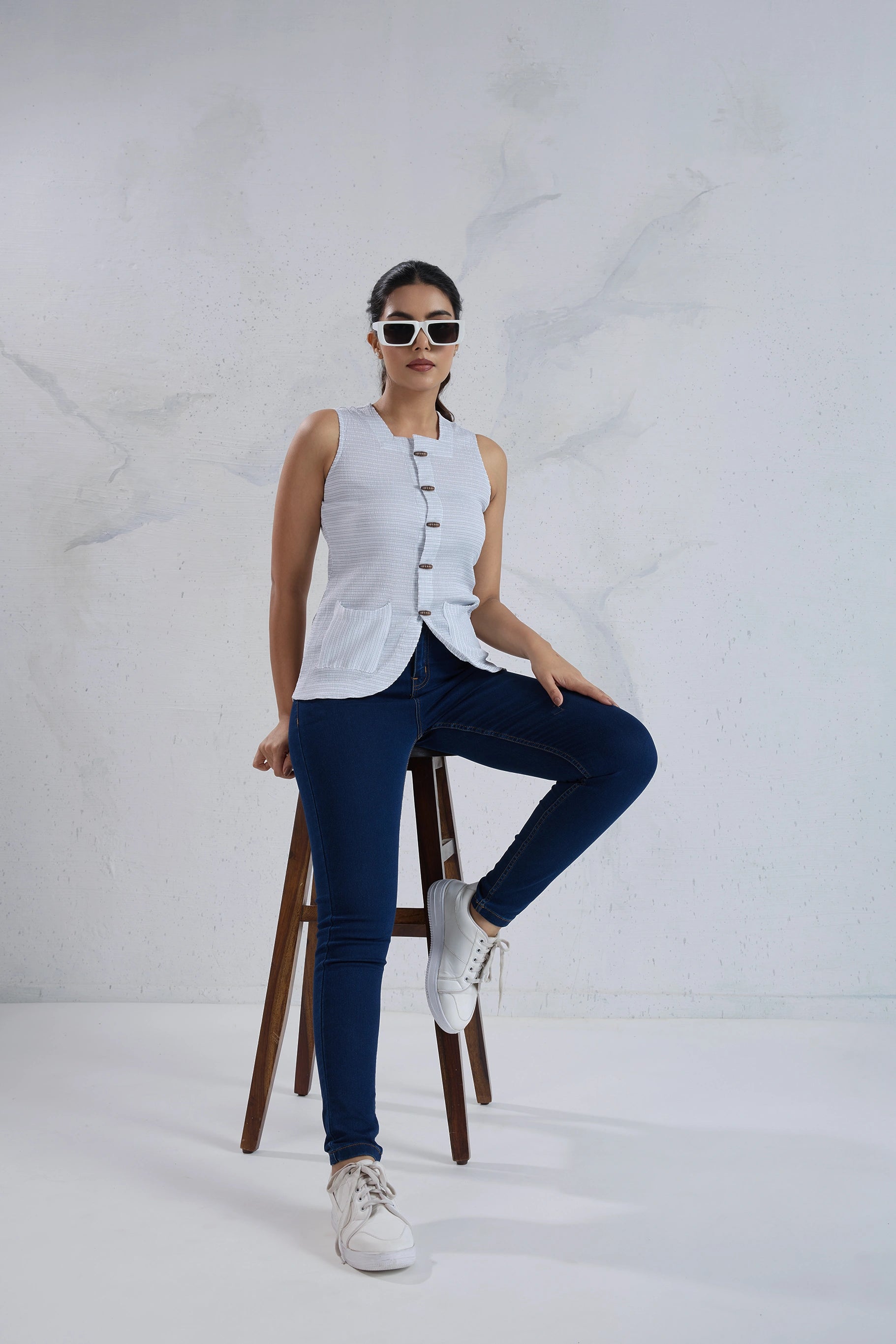 Woman sitting on a stool wearing a white sleeveless top and blue jeans against a white background