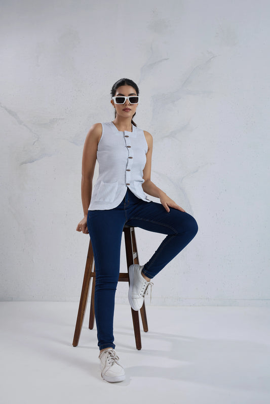 Woman sitting on a stool wearing a white sleeveless top and blue jeans against a white background