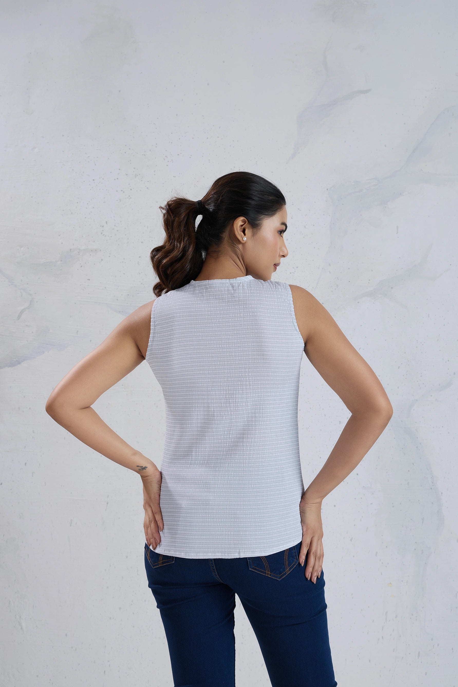 Woman wearing a sleeveless white top and blue jeans against a light gray background