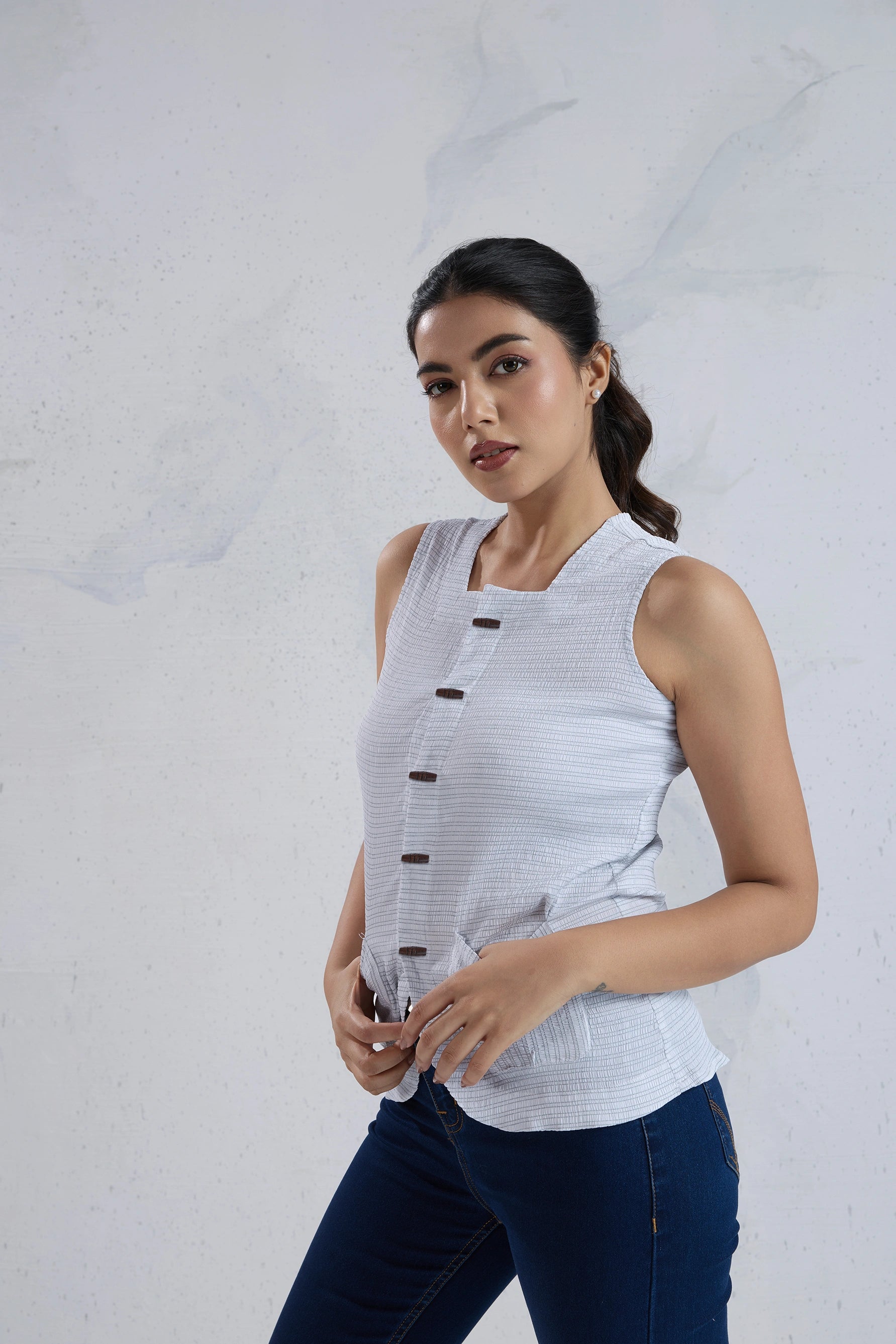 Woman wearing a sleeveless white top with black buttons against a light gray background