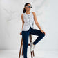 Woman sitting on a stool wearing a white sleeveless shirt, dark jeans, and white sneakers against a white background.