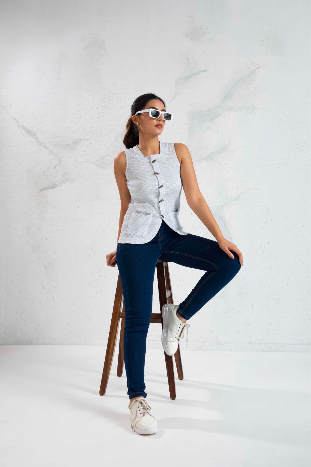 Woman sitting on a stool wearing a white sleeveless shirt, dark jeans, and white sneakers against a white background.
